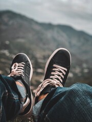 A black pair of sneakers with white laces and with blue jeans is waving in front of the mountains