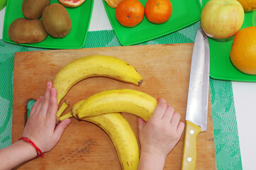 Children's hands prepare bananas for slicing on the kitchen board. 