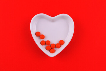 Small red pills in a white plate in the shape of a heart on a red background