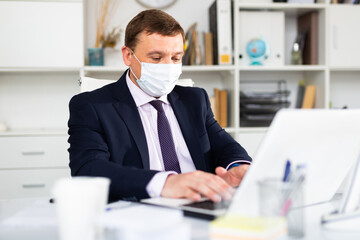 Focused businessman in disposable face mask working on laptop in office. Necessary precautions during COVID 19 pandemic