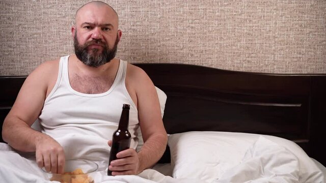 A plump bearded man watches TV, drinks beer, eats chips, sitting at home in bed. food falls on the shirt copy space