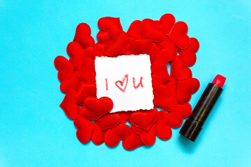 On the red hearts is a white square with a declaration of love written in lipstick, which lies next to it. Blue background. Minimal Valentine's Day greeting card with copy space.