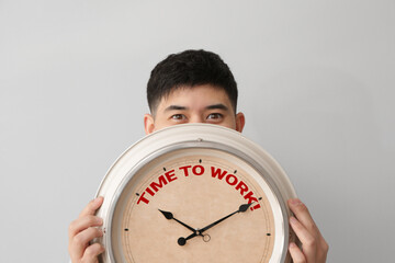 Young Asian man holding clock with text TIME TO WORK on light background