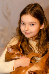 A child, 7-year-old girl with long blonde hair playing with a cat at home on the couch. Pets and children concept. Red cat breed Abyssinian.