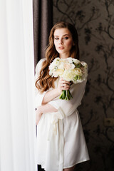 beautiful woman in a white robe with a wedding bouquet of flowers at the window