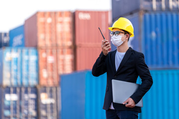 Asian male wear a mask use a radio to command logistic warehouse foreman checking list container boxes. industry import export shipping business, During the spread of the coronavirus or covid-19.