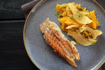 Cod fish with fennel vegetable and orange slices on a blue plate and a dark table, high angle view from above