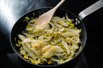 Fennel slices and leek in a black frying pan on the stove, cooking vegetarian with healthy...