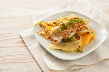 Cod fish fillet on sauteed fennel vegetable with orange slices and leek served on a white plate and napkin, light wooden table with copy space, selected focus