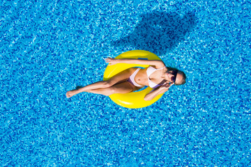 Beautiful woman in swimming pool aerial top view from above. Young girl in bikini relaxes and swims on inflatable ring donut and has fun in water on family vacation, tropical holiday resort