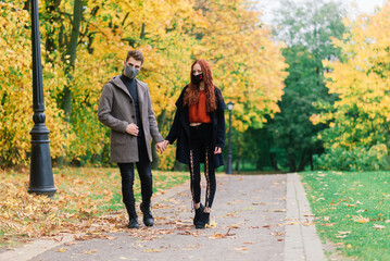 Young red haired woman puts on face mask while walking with young man in autumn park.