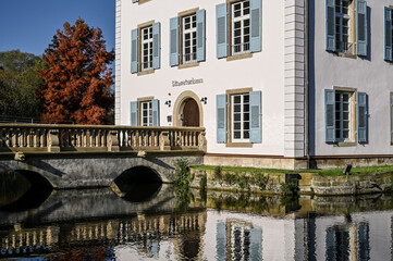 A baroque house, called Trappenseeschlösschen, surrounded by Lake Trappensee in Heilbronn, Germany during autumn. A pedestrian bridge leads to the house.