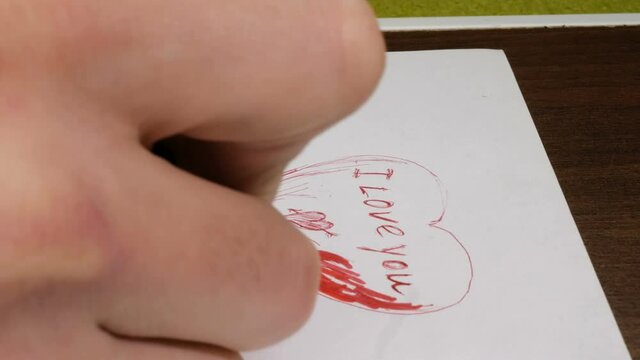 Close-up of a young man's hand he writes on paper the words I love you with a red pen.