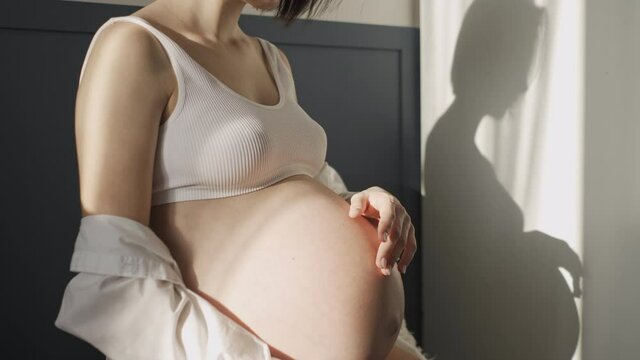 Young pregnant girl in white underwear and white shirt near a white wall in the sun
