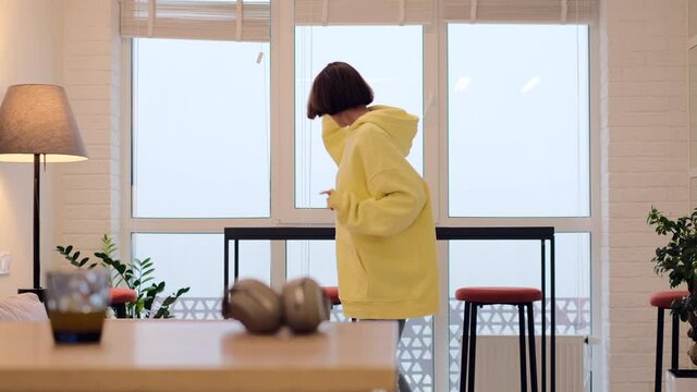 Young woman at home in the living room in the morning in a giant yellow sweater dances by the window, enjoys the time alone, positively attunes to the day, cloudy and gloomy outside the window   