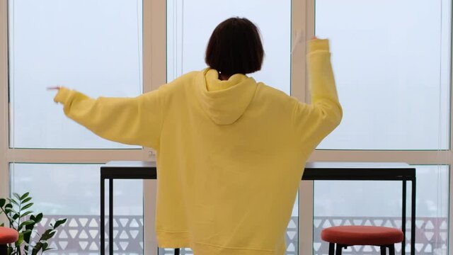 Young woman at home in the living room in the morning in a giant yellow sweater dances by the window, enjoys the time alone, positively attunes to the day, cloudy and gloomy outside the window     