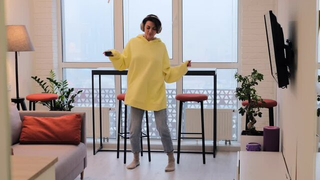 Young woman at home in the living room in the morning in a giant yellow sweater dances by the window, enjoys the time alone, positively attunes to the day, cloudy and gloomy outside the window   