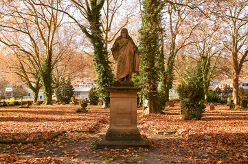 A statue of Jesus is standing in a graveyard that is covered with brown leaves. The words on the socket "Friede sei mit euch" means "peace be with you".