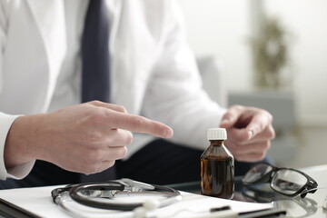 The doctor points with his hands to the bottle with the vaccine which is on the glass table