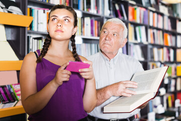 Teen girl with phone in bookstore and grandfather choosing book