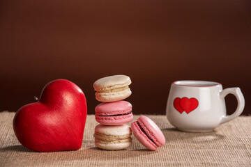 4 French pink and vanilla macarons, one red love heart and one coffee cup with a red heart painted on it, placed on a beige fabric and a chocolate background 