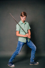 A teenage boy in a green T-shirt and jeans holds a medieval sword in his hands. Studio photo on a gray background.