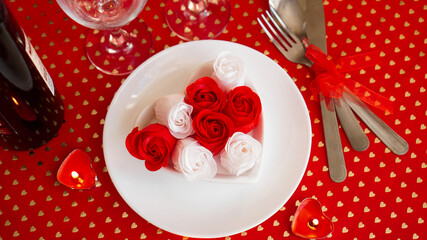 A white plate with wine, a knife and fork on a bright red background. Red and white roses decoration. Table setting for Valentines Day