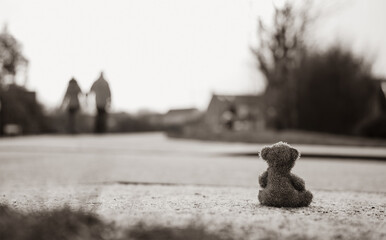 Rear view Lonely Teddy bear doll sitting alone on hailstones with blurry couple walking on footpath.lost brown bear toy looking out on the road, International missing children's day