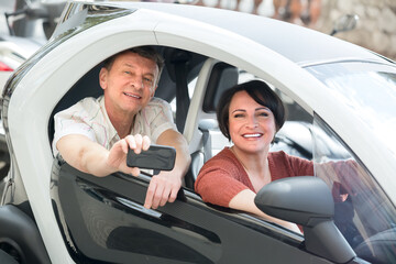Cheerful smiling mature couple driving twizy electric outside at summer. Focus on woman.