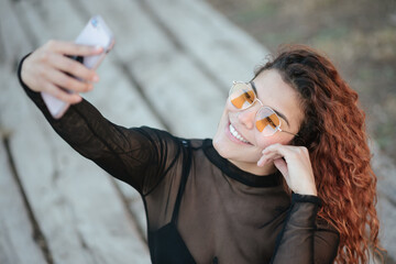 Pretty latin woman with sunglasses doing a self portrait with her smarphone sitting on a wooden path in the countryside, outdoors