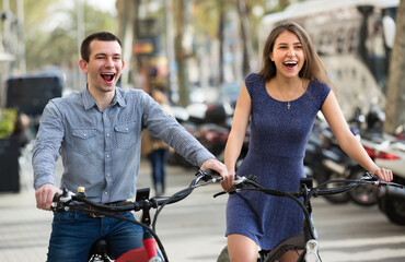 Obraz premium Cheerful young couple riding on electric bikes outside and laughing. Selective focus.