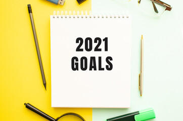 Notebook with text 2021 GOALS on office table with office supplies. Yellow color background.