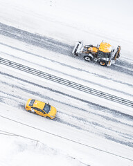 Winter road, on a snow-covered street, a yellow tractor and a taxi are moving in the opposite direction. View from above. Blizzard in the city.