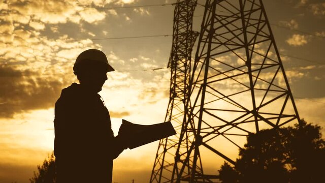silhouette of an electrician engineer working next to an electrical support tower sunset energy. electricity high voltage tower support. electrical engineer inspect voltage electricity pylon