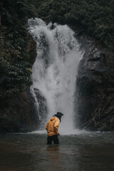 Fototapeta na wymiar An asian male traveler enjoying the beauty of rainforest waterfall.A male explorer in the waterfall pool.Camping and hiking lifestyle.Into the wild.