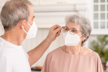 Senior man putting on surgical mask on his elderly wife ill woman during the coronavirus epidemic