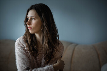 Young, attractive, caucasian white woman with brown, curly hair sitting on a sofa on a cold, rainy winter day and looking outside the window. Sad, lost, lonely feeling context