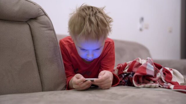 Little boy is lying on couch with smartphone in hands and playing mobile game or using social networks. Modern problem of dependence of kids on gadgets and Internet from early age.
