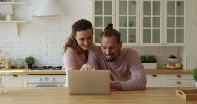 Young couple spending weekend time in kitchen with laptop, wife and husband looks at computer screen discuss purchase, booking hotel planning vacation, enjoy comfort e-commerce buying on-line concept