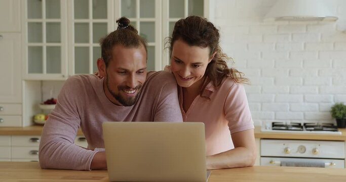 30s boyfriend girlfriend discuss purchase on internet standing in domestic kitchen looking at laptop screen choose goods online, couple making food delivery order. E-commerce satisfied clients concept