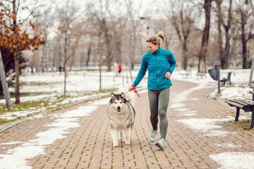 Sportswoman running with her dog in a park on cold winter day. Pets, winter fitness, runner,...