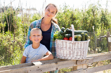 Harvesting season. Portrait of young woman with son posing with harvest of vegetables on sunny garden