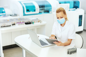 Fototapeta na wymiar Attractive blond lab assistant in coat with sterile surgical mask on sitting in laboratory, entering test results on tablet. Research for cure for covid 19 concept.