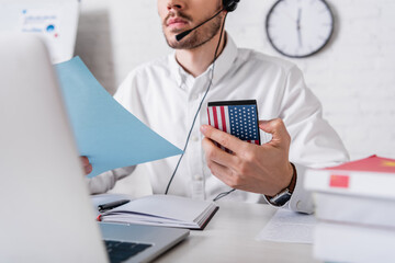 partial view of interpreter in headset holding paper and digital translator with usa flag emblem near laptop on blurred foreground