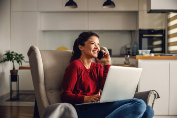 Smiling young woman sitting at home and having phone conversation with her boss. In lap she is holding laptop. Remote business during corona virus concept.