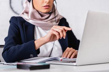 cropped view of arabian interpreter pointing with finger near laptop and digital translator on...