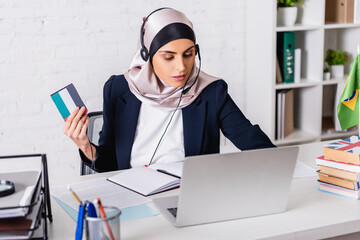 arabian interpreter in headset holding digital translator while working on laptop near dictionaries, blurred foreground
