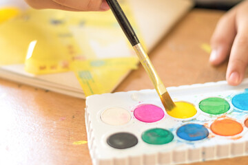 Colorful water color cakes in palette with paint brush on yellow colour in kid's hand to do art craft on notebook cover in class. School student doing art home work kraft on wooden table