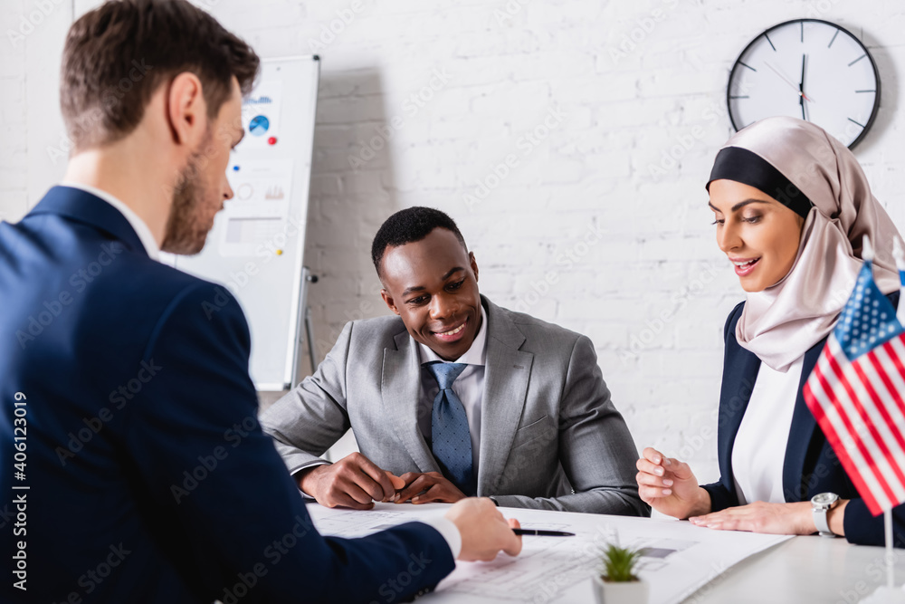 Wall mural cheerful african american and arabian business partners looking at blueprint near translator on blurred foreground