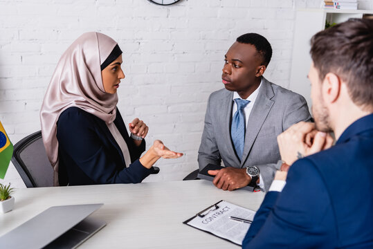 arabian businesswoman pointing with hand during meeting with multicultural business partners near contract, blurred foreground